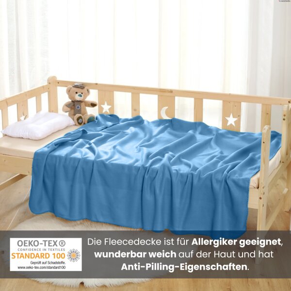 Eine blaue Kuscheldecke aus Fleece liegt zusammen mit einem Teddy und einem Kissen auf einem Kinderbett aus hellem Holz.