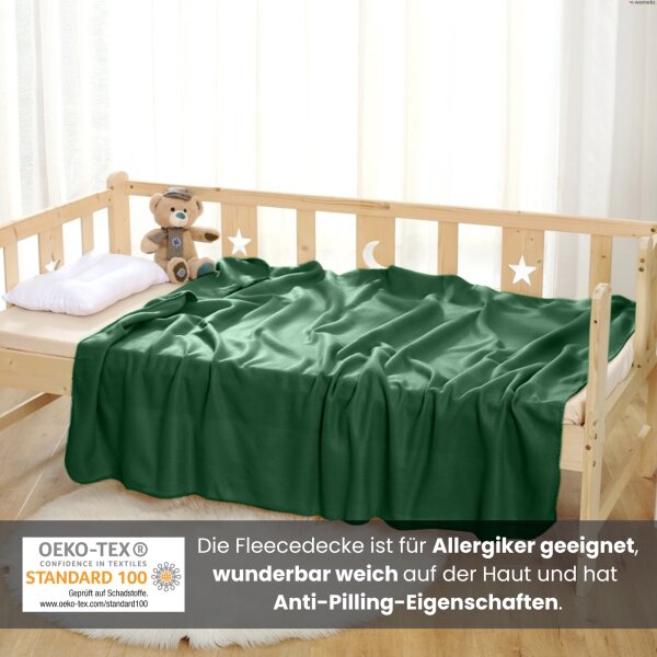 Eine dunkelgrüne Kuscheldecke aus Fleece liegt zusammen mit einem Stofftier und einem Kissen auf einem Kinderbett.