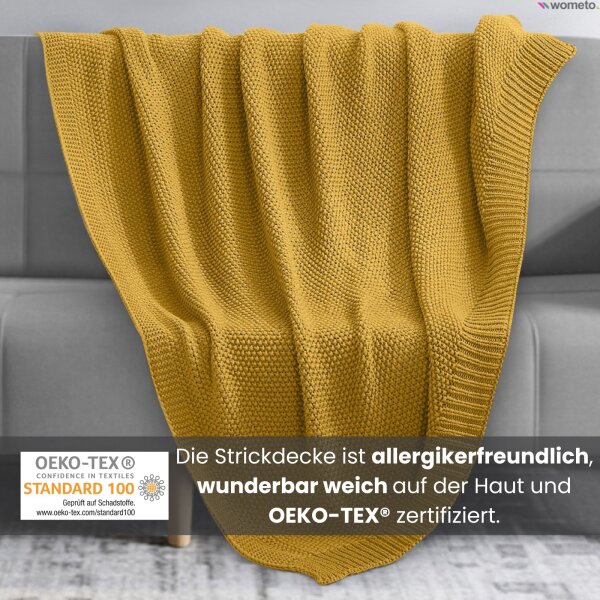 Eine gelbe Strickdecke liegt auf einem grauen Sofa. Darunter der Hinweis, die Kuscheldecke ist OEKO-TEX zertifiziert.