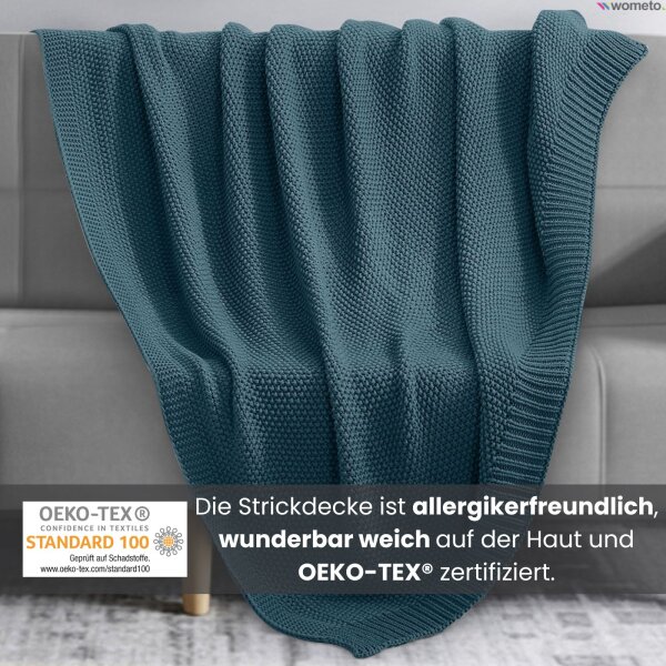 Eine türkise Strickdecke liegt auf einer grauen Couch. Darunter der Hinweis, die Kuscheldecke ist allergikerfreundlich.