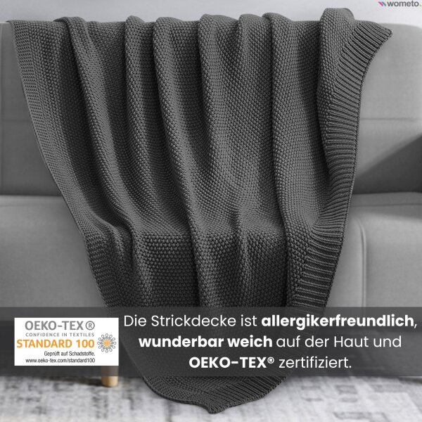 Eine dunkelgraue Strickdecke liegt auf einer grauen Couch. Darunter der Hinweis, die Kuscheldecke ist allergikerfreundlich.