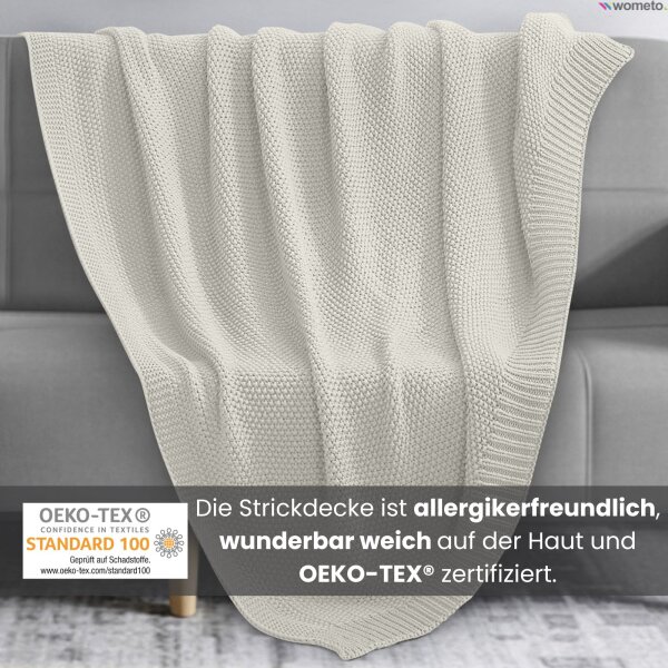 Eine weiße Strickdecke liegt auf der grauen Couch. Darunter der Hinweis, die kuschelige Decke ist OEKO-TEX zertifiziert.