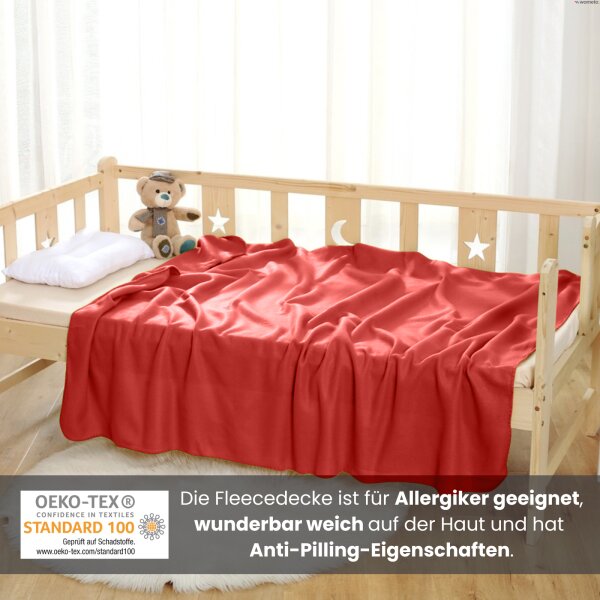 Eine rote Kuscheldecke aus Fleece liegt zusammen mit einem Teddybär und einem Kissen auf dem Kinderbett.